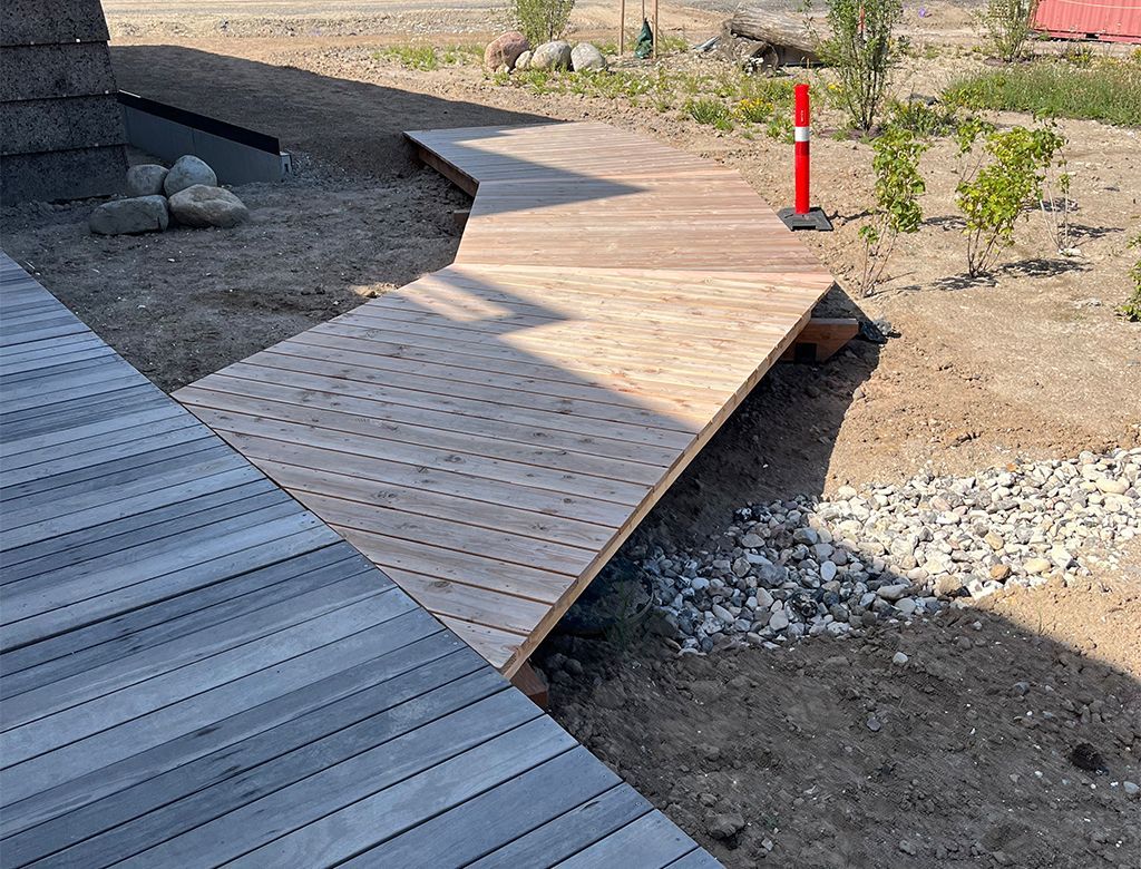 Træsti over jord og grus i solrige udendørs omgivelser.