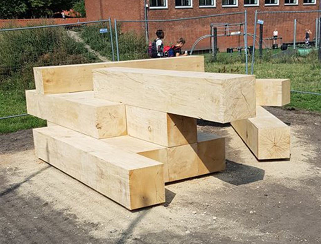 Legepladsstruktur af træ lavet af stablede, lyse træklodser på en jordoverflade med børn i baggrunden.