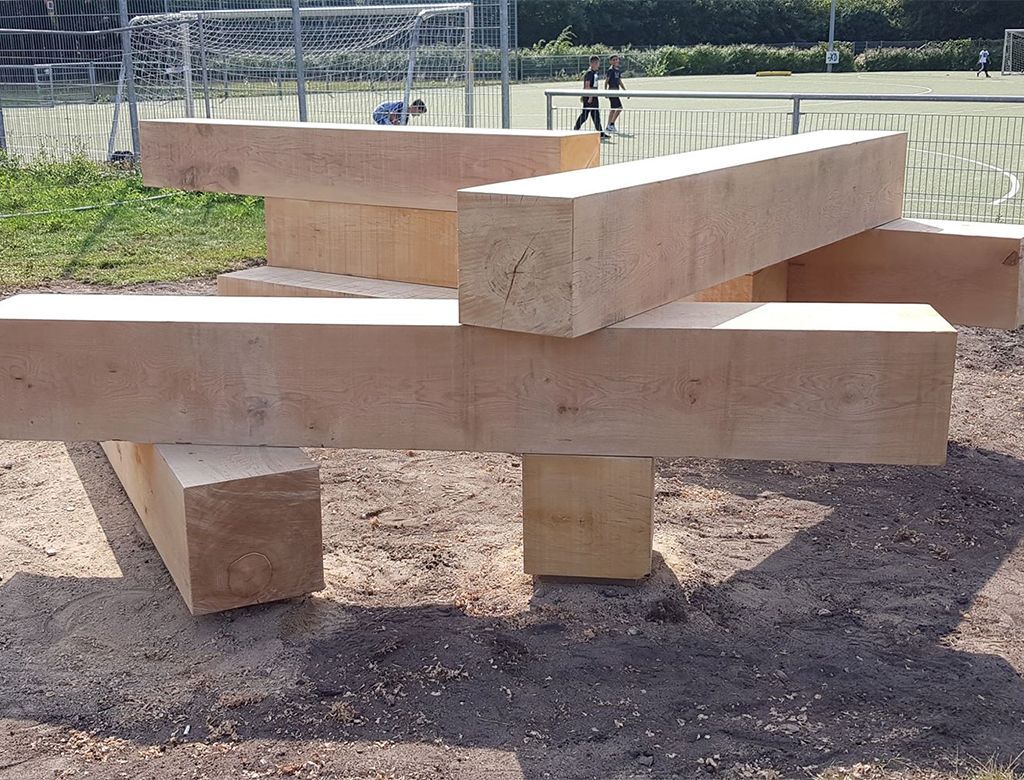 Træbjælker arrangeret i en legende, abstrakt struktur på en sandet overflade nær en græsklædt sportsbane.