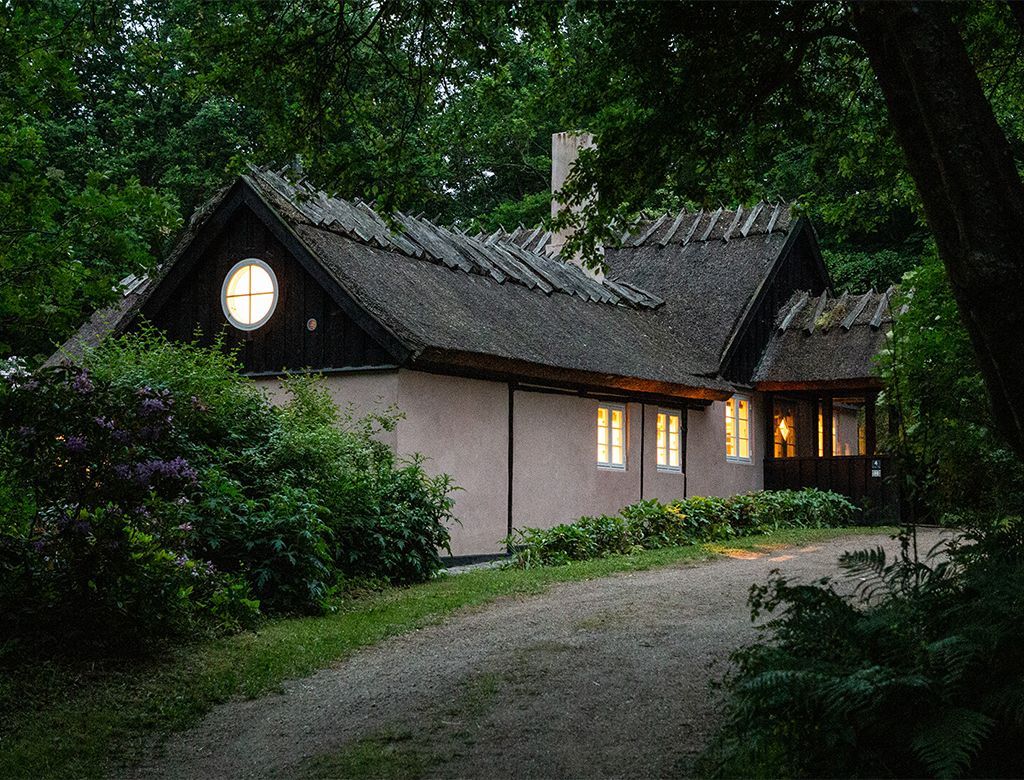 Hytte med stråtag og lysende vinduer på en grussti omgivet af træer og buske.