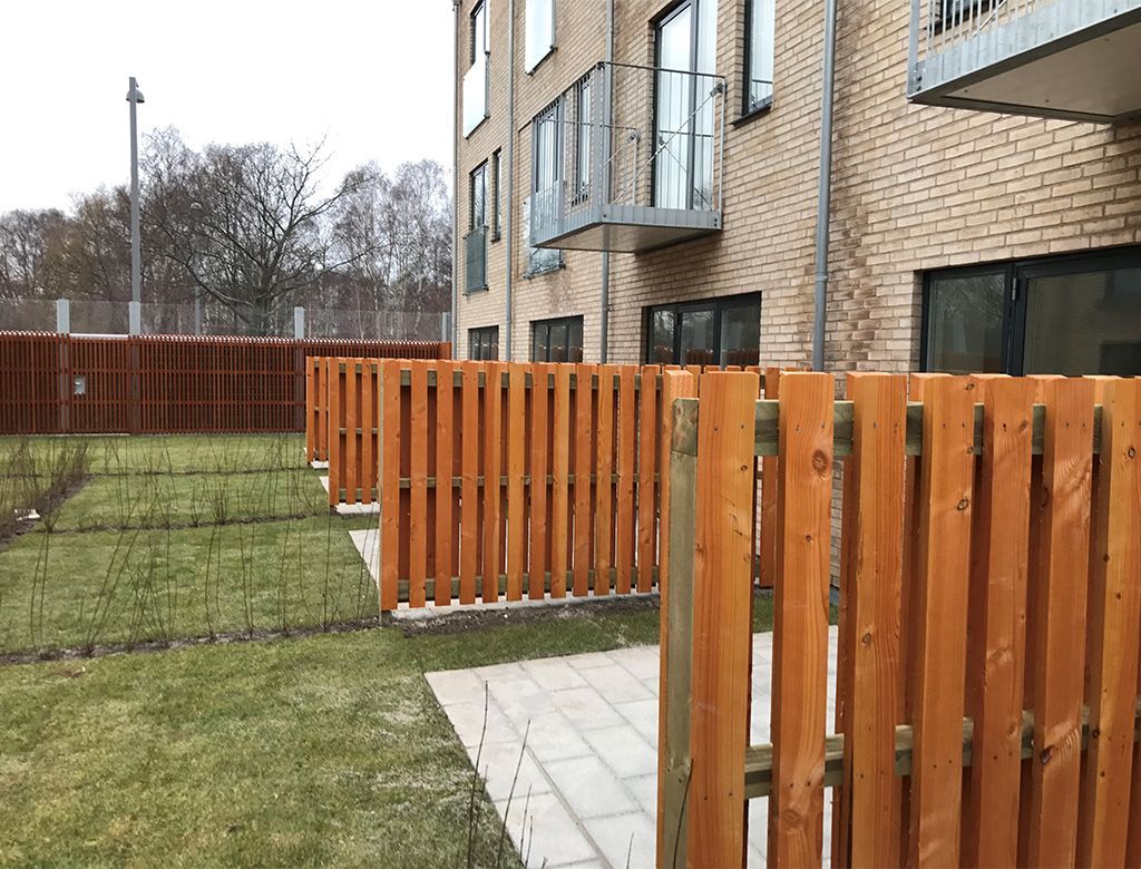 Træhegn foran en murstensbygning, grøn græsplæne og grå belægningssten.