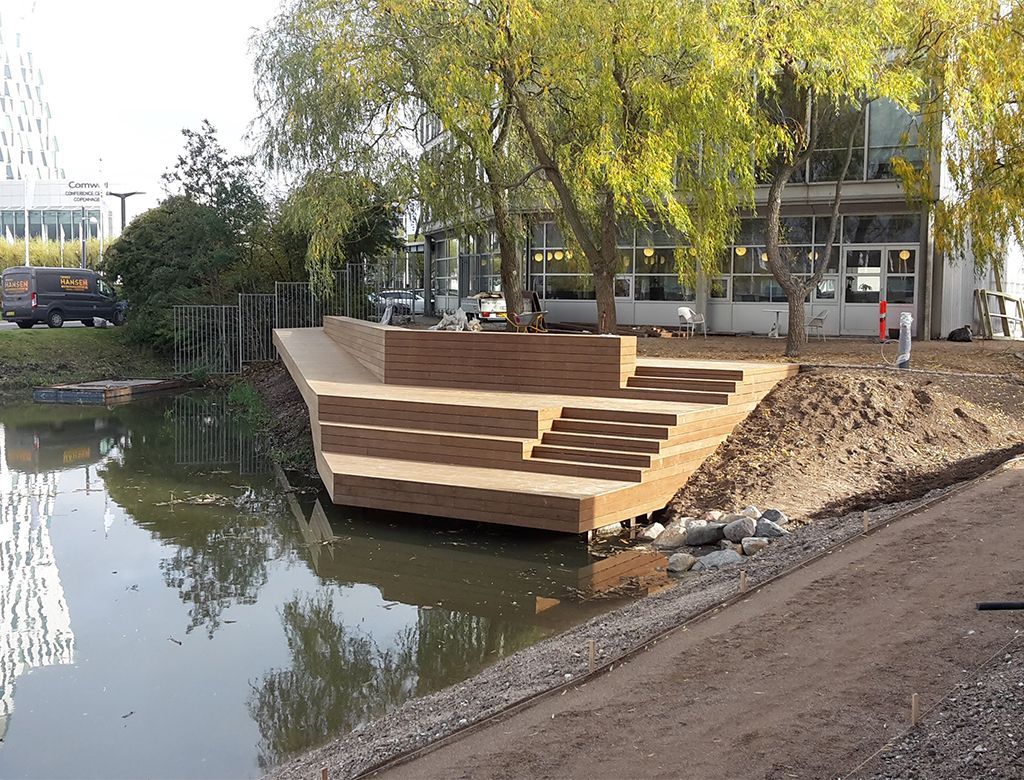 Trætrappe og platform bygget ved vandkanten, ved siden af en bygning og træer.