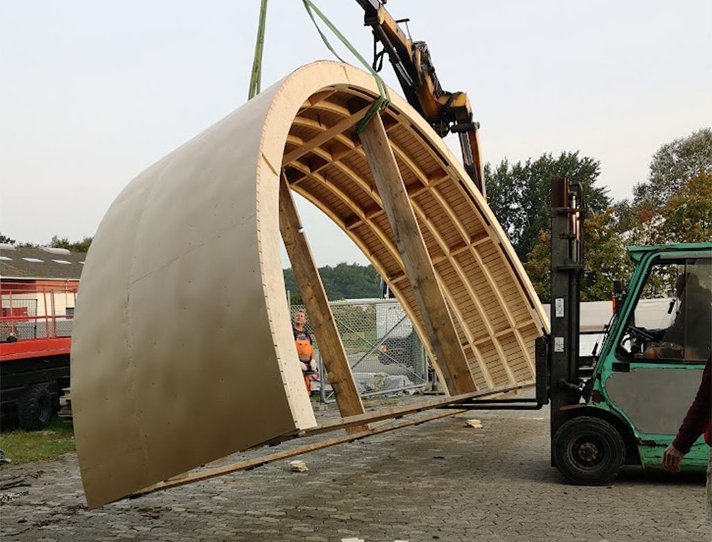 En stor, buet arkitektonisk sektion løftes udendørs med en kran og gaffeltruck.