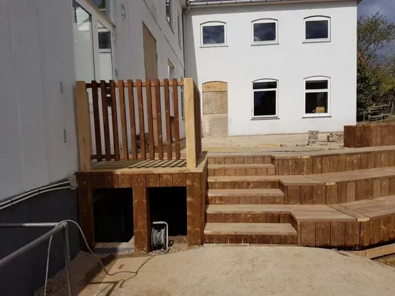 Træramme af et hus under opførelse, blå himmel, byggematerialer og et delvist færdiggjort hus i baggrunden.