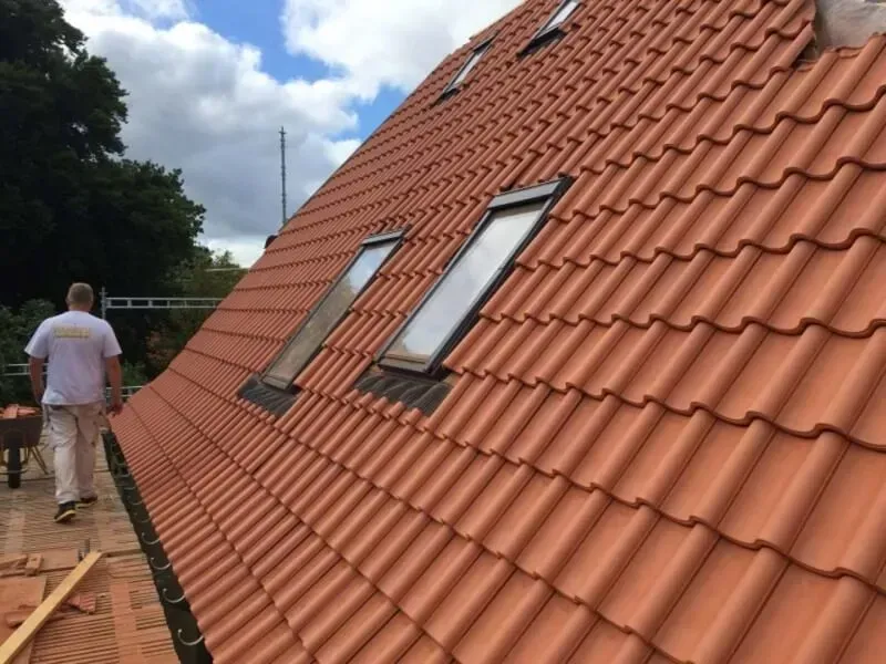 Træramme af et hus under opførelse, blå himmel, byggematerialer og et delvist færdiggjort hus i baggrunden.