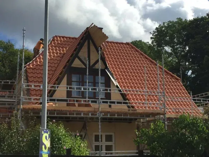 Træramme af et hus under opførelse, blå himmel, byggematerialer og et delvist færdiggjort hus i baggrunden.