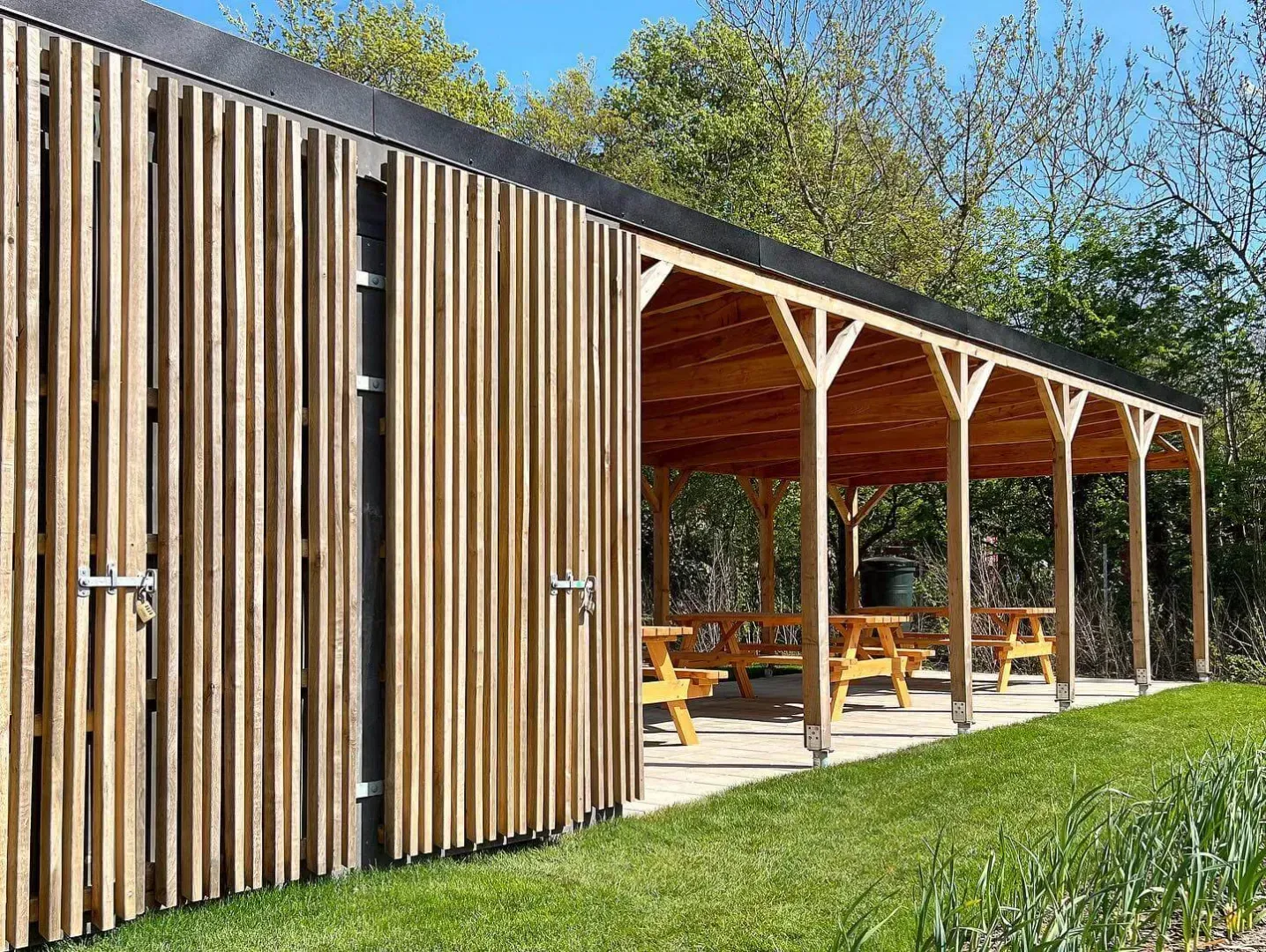 Træly med picnicborde; solrige udendørs omgivelser.