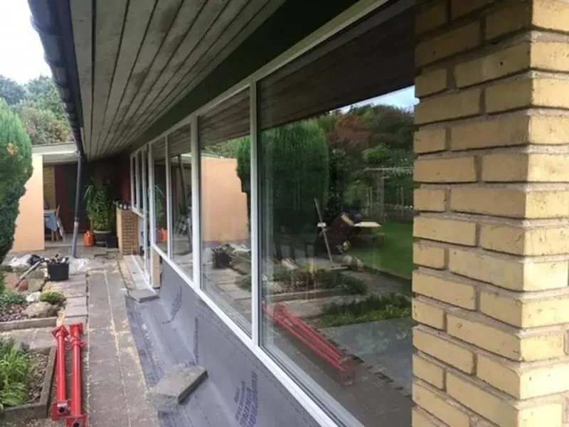 Træramme af et hus under opførelse, blå himmel, byggematerialer og et delvist færdiggjort hus i baggrunden.