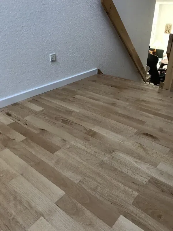 Træramme af et hus under opførelse, blå himmel, byggematerialer og et delvist færdiggjort hus i baggrunden.