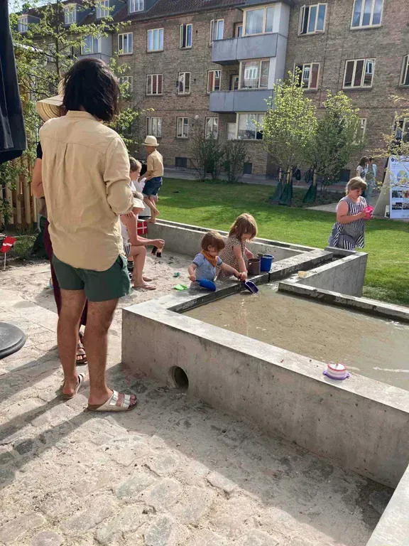 Børn leger udendørs i et betonvandsspejl. En voksen står i nærheden og ser på. Murstensbygning i baggrunden.