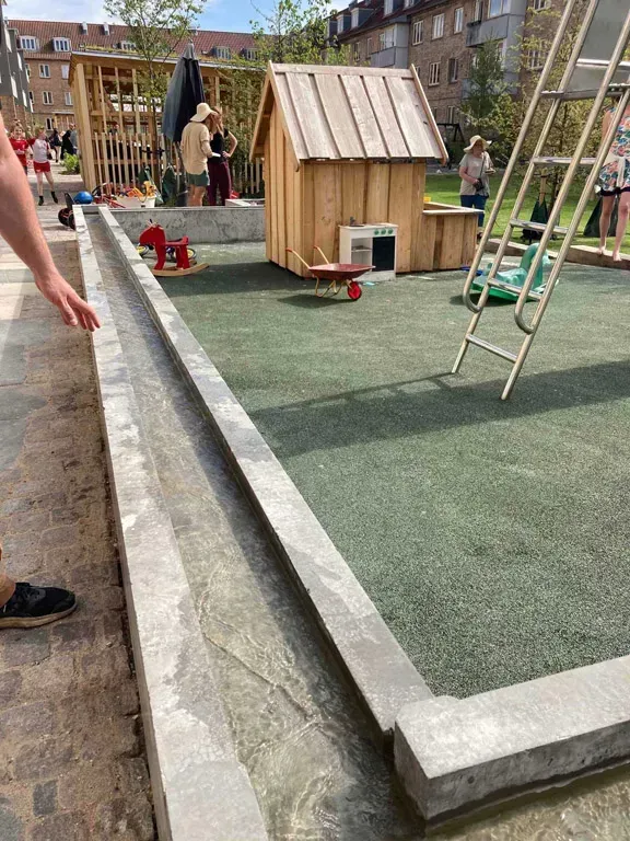 En person rører ved en betonvandkanal på en legeplads med et lille træhus og en stige.