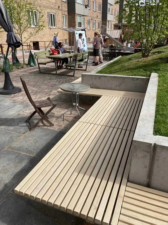 Udendørs siddeområde med træbænke, et lille bord og folk, der nyder pladsen.