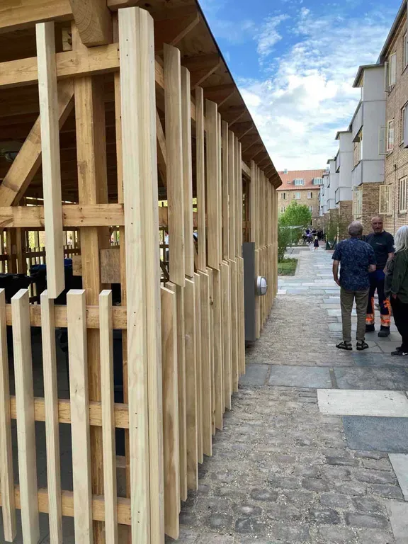 Træstruktur med lodrette lameller, mennesker stående på en brostensbelagt sti. Bygninger i baggrunden.