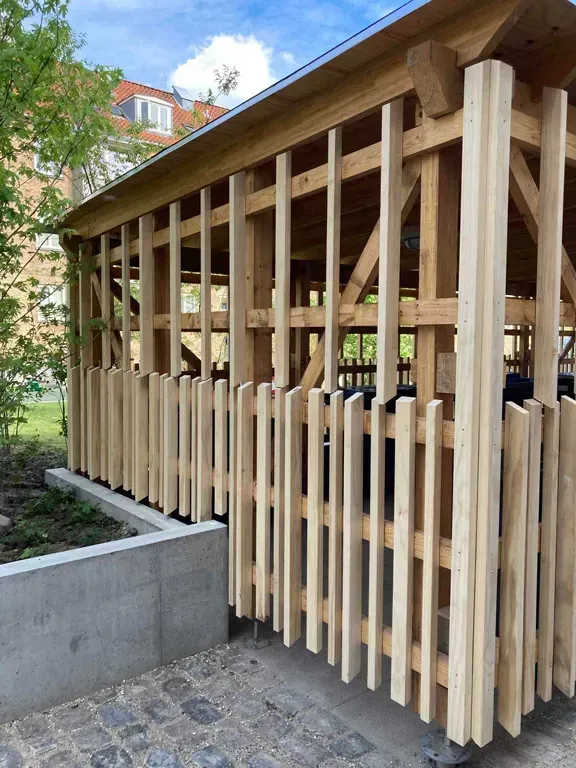 Træpavillon med lodrette lameller, beliggende i en park med en murstenssti og betonfundament.