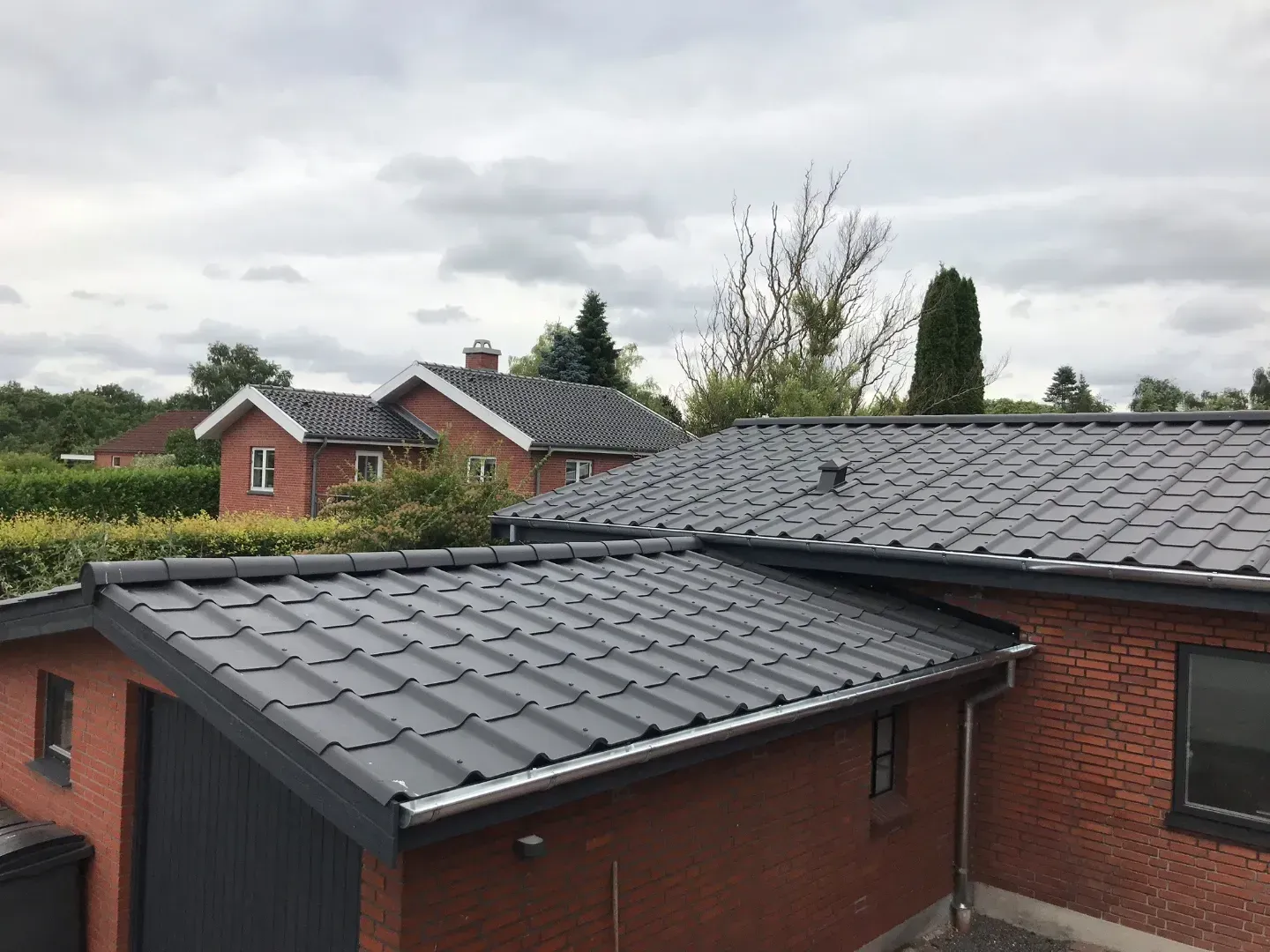 Træramme af et hus under opførelse, blå himmel, byggematerialer og et delvist færdiggjort hus i baggrunden.