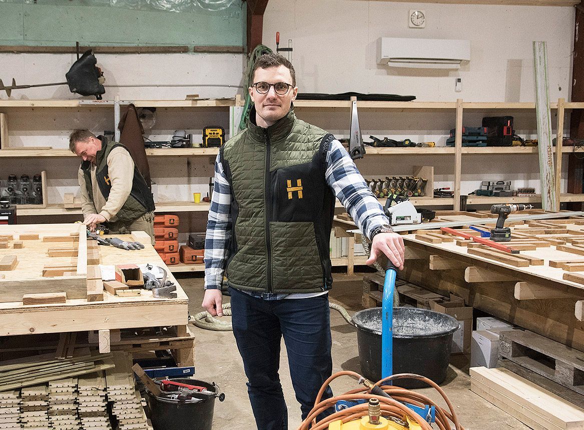 En mand i vest står i et værksted og holder en blå dolly med et orange snor, mens en anden person arbejder i baggrunden.