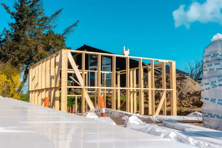 Træramme af et hus under opførelse, blå himmel, byggematerialer og et delvist færdiggjort hus i baggrunden.