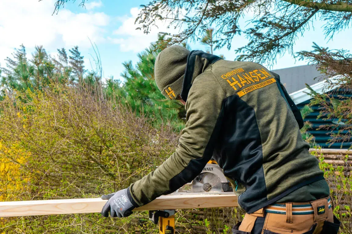 Tømrer i grønt arbejdstøj saver træ udenfor i klart dagslys.