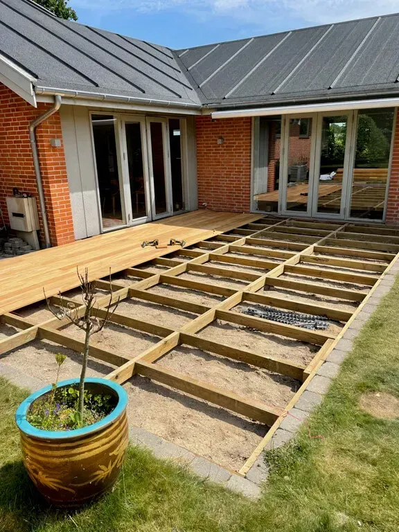 En træterrasse under opførelse i en baghave med et murstenshus og en potteplante i udsigt.
