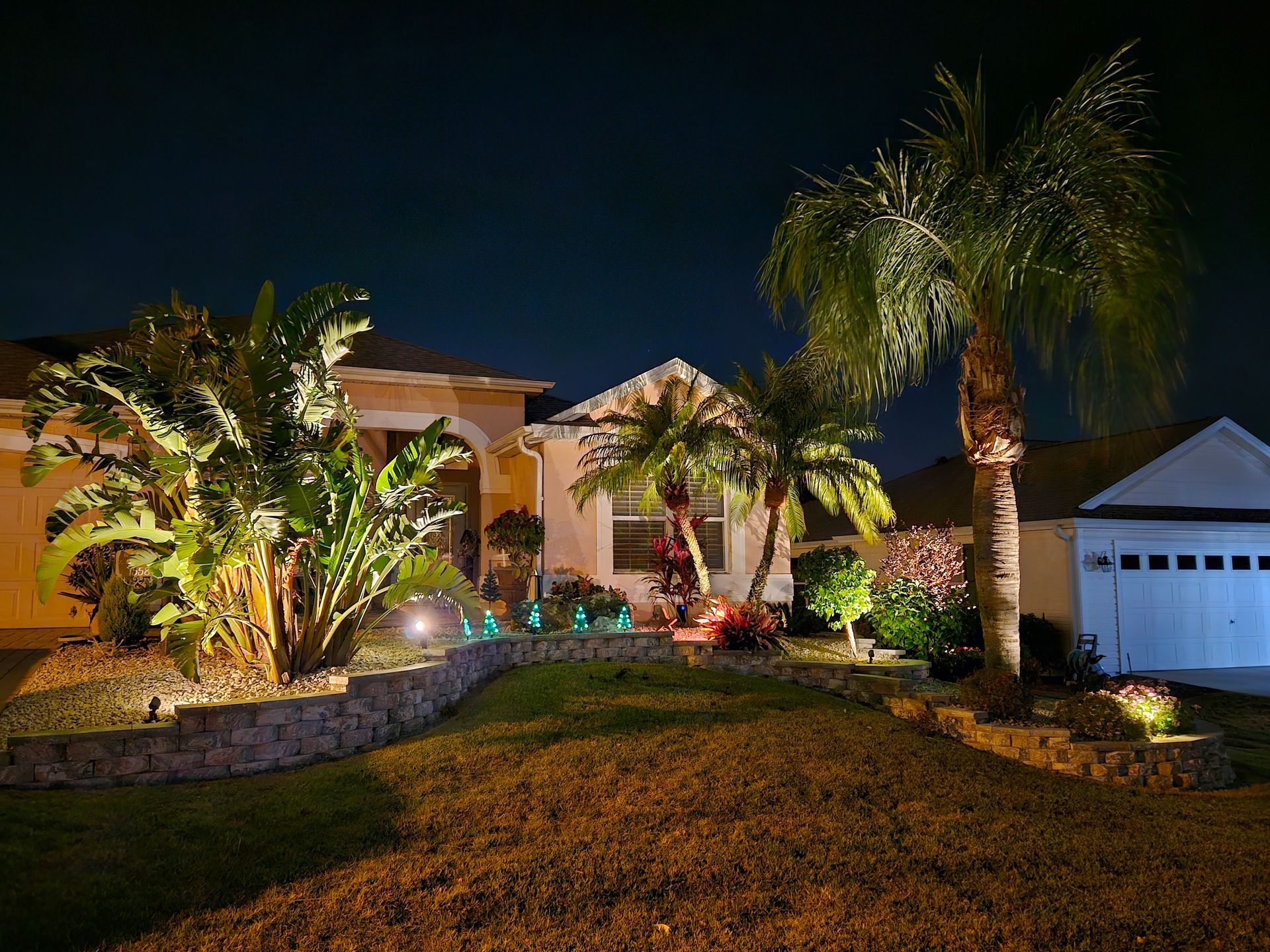 Night view of a house with illuminated landscaping, including palm trees, a retaining wall, and a grassy lawn.