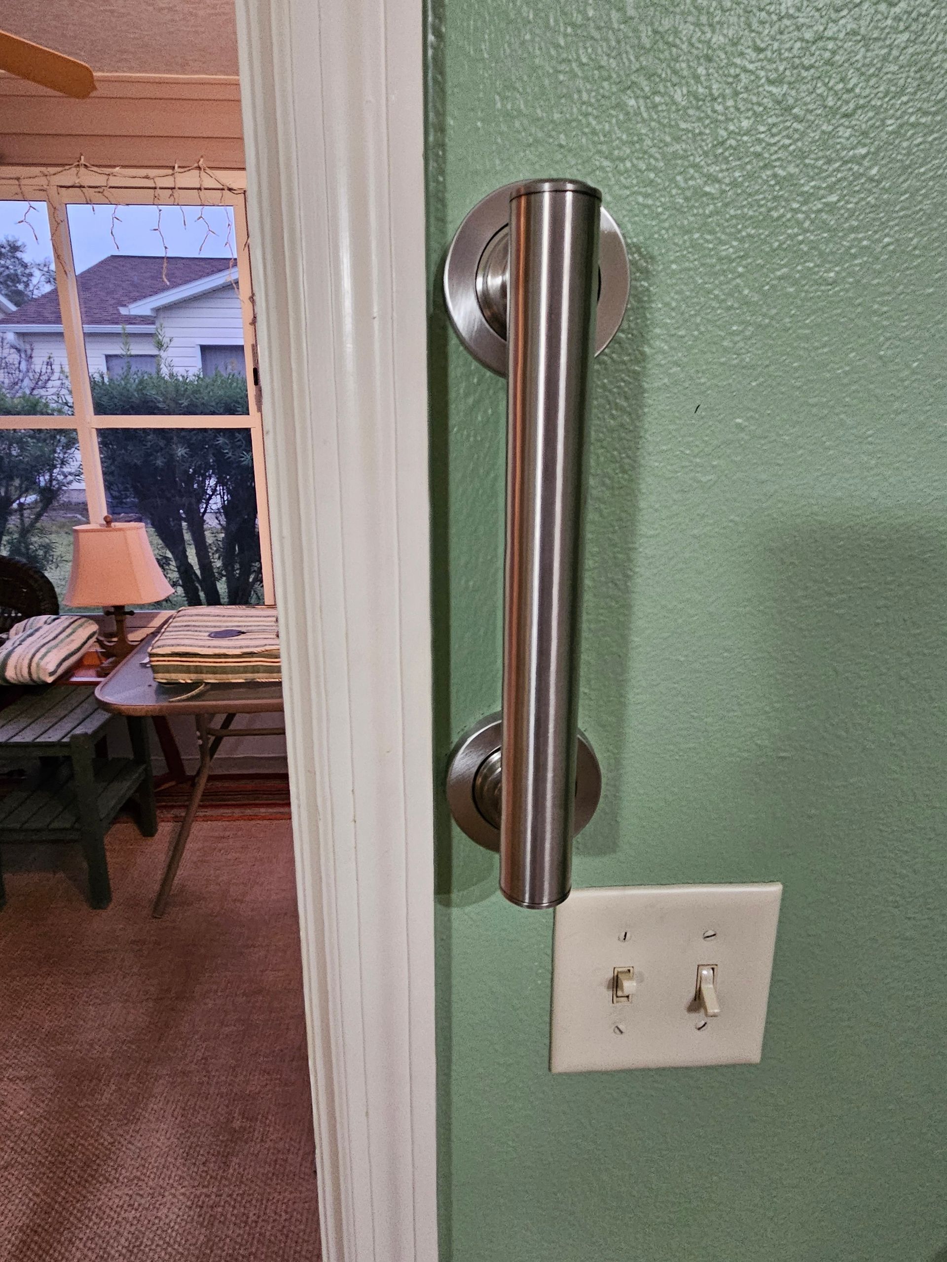 Stainless steel vertical grab bar mounted on a light green wall next to a white light switch.