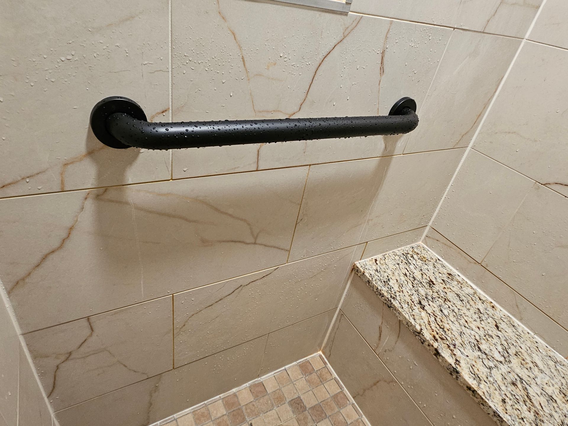 Black grab bar on a shower wall with beige tile, over a granite step.