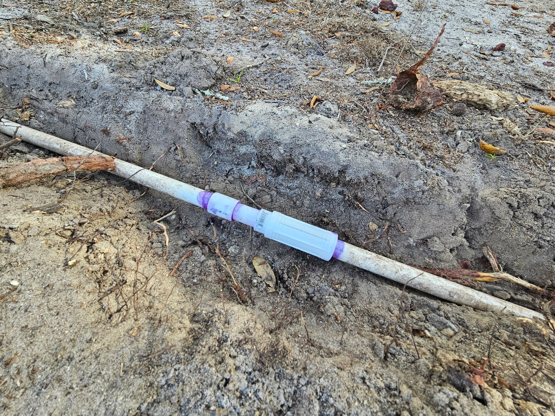 White pipe with a blue fitting lies in a sandy trench, surrounded by debris.