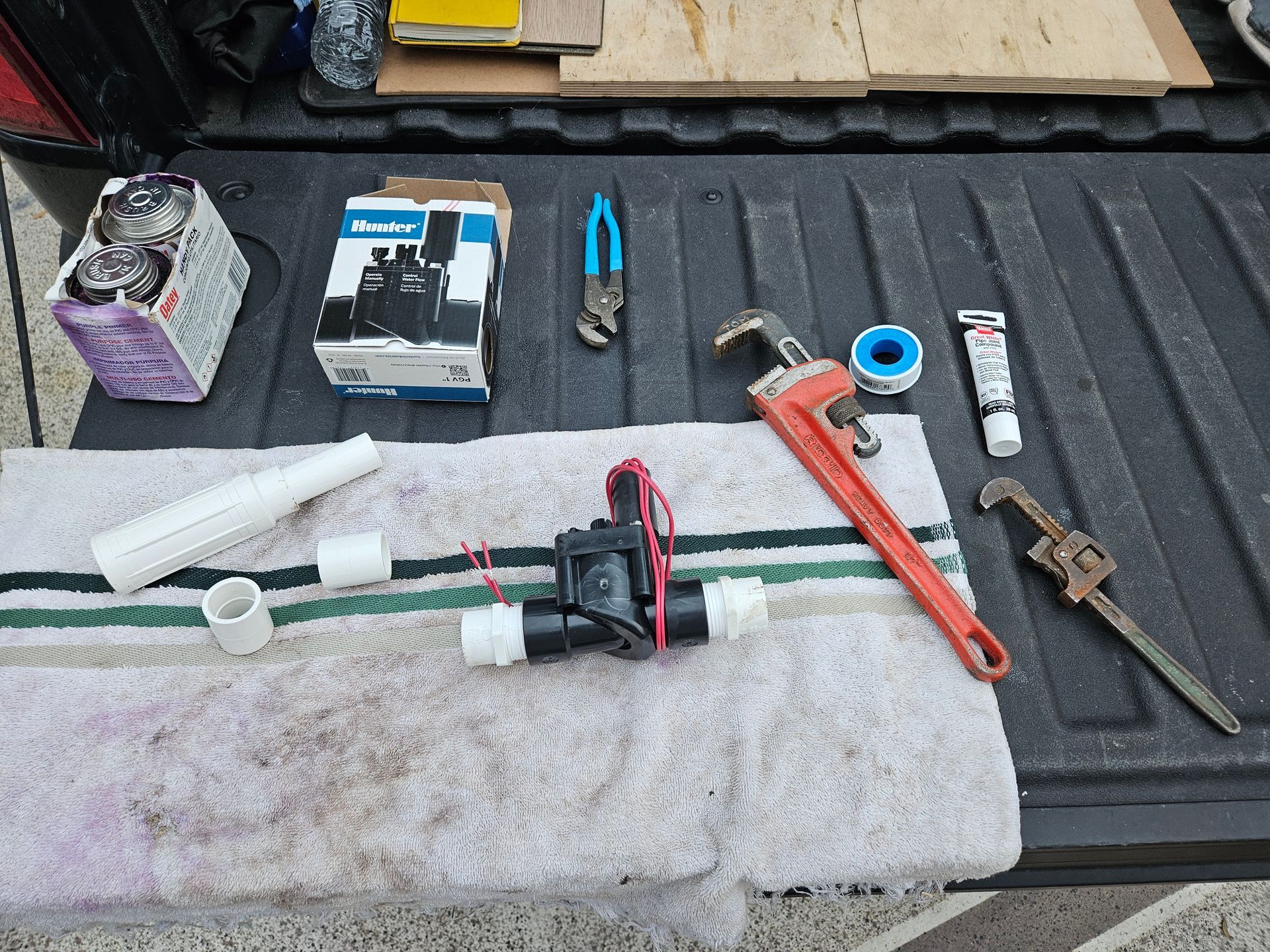 Plumbing parts and tools laid out on a truck bed: PVC pipe, valve, wrenches, pliers, tape, and a sealant.