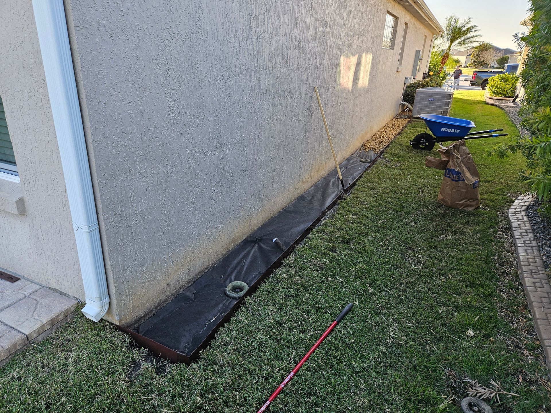 Black landscape fabric against a beige house. Green grass surrounds, with a blue wheelbarrow in the background.