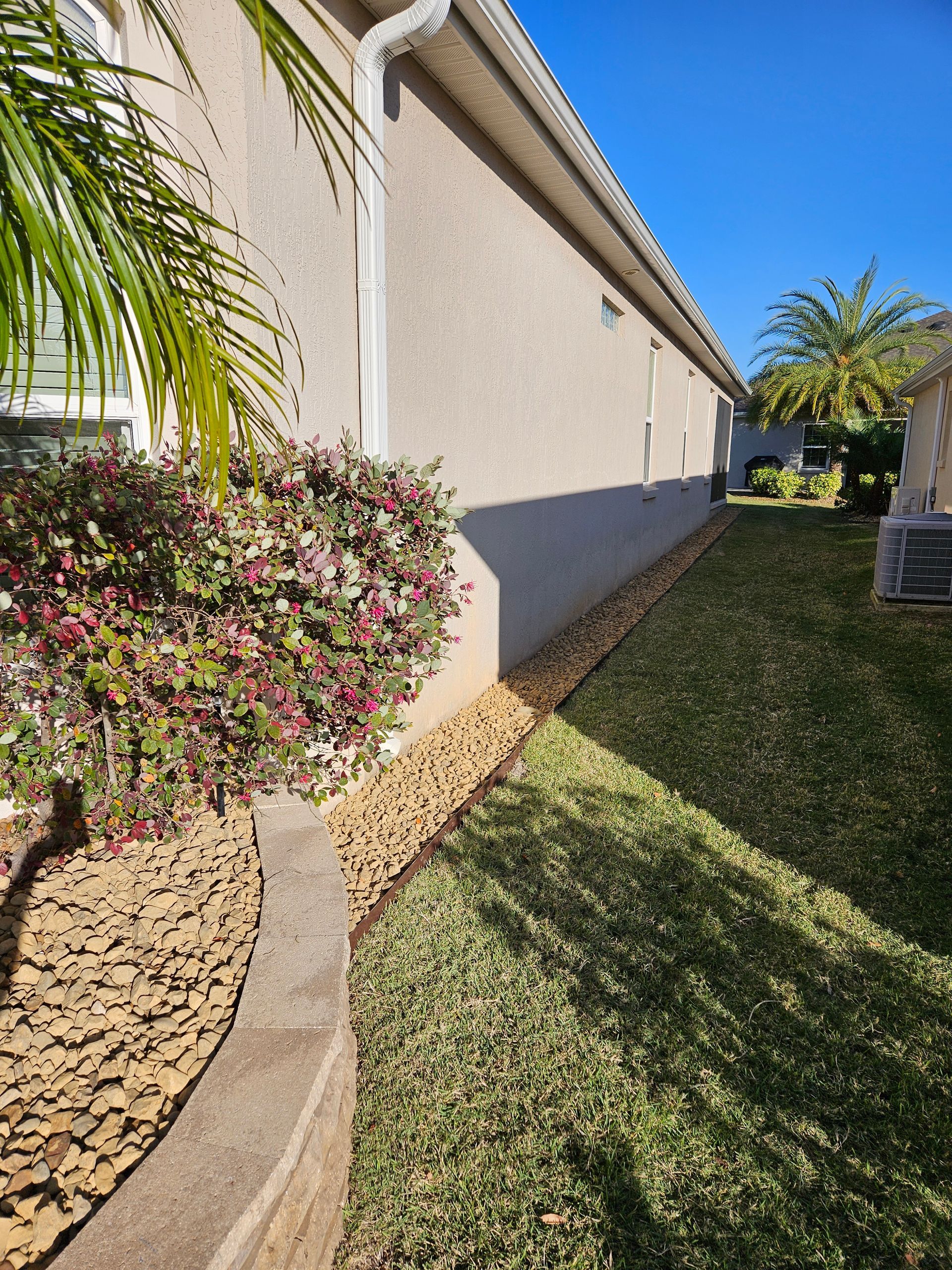 Beige house exterior with a rock bed, lush green grass, and clear blue sky.