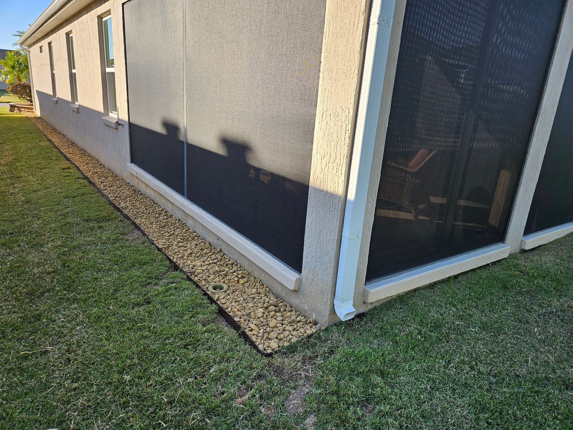 A house with black screens and tan walls. Gravel borders the side along the grass.