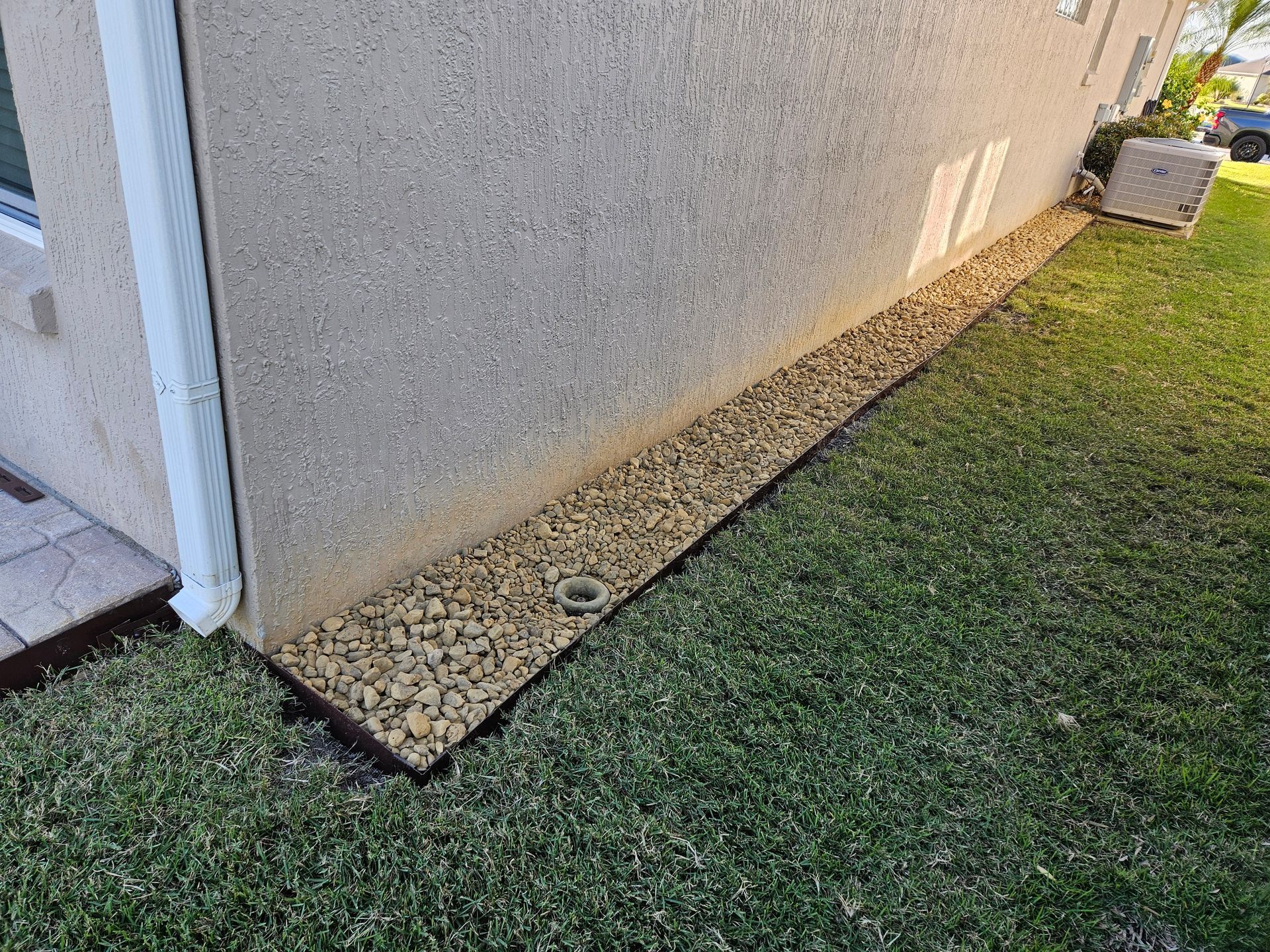 Gravel border along a light-colored stucco wall and green lawn, near a building.