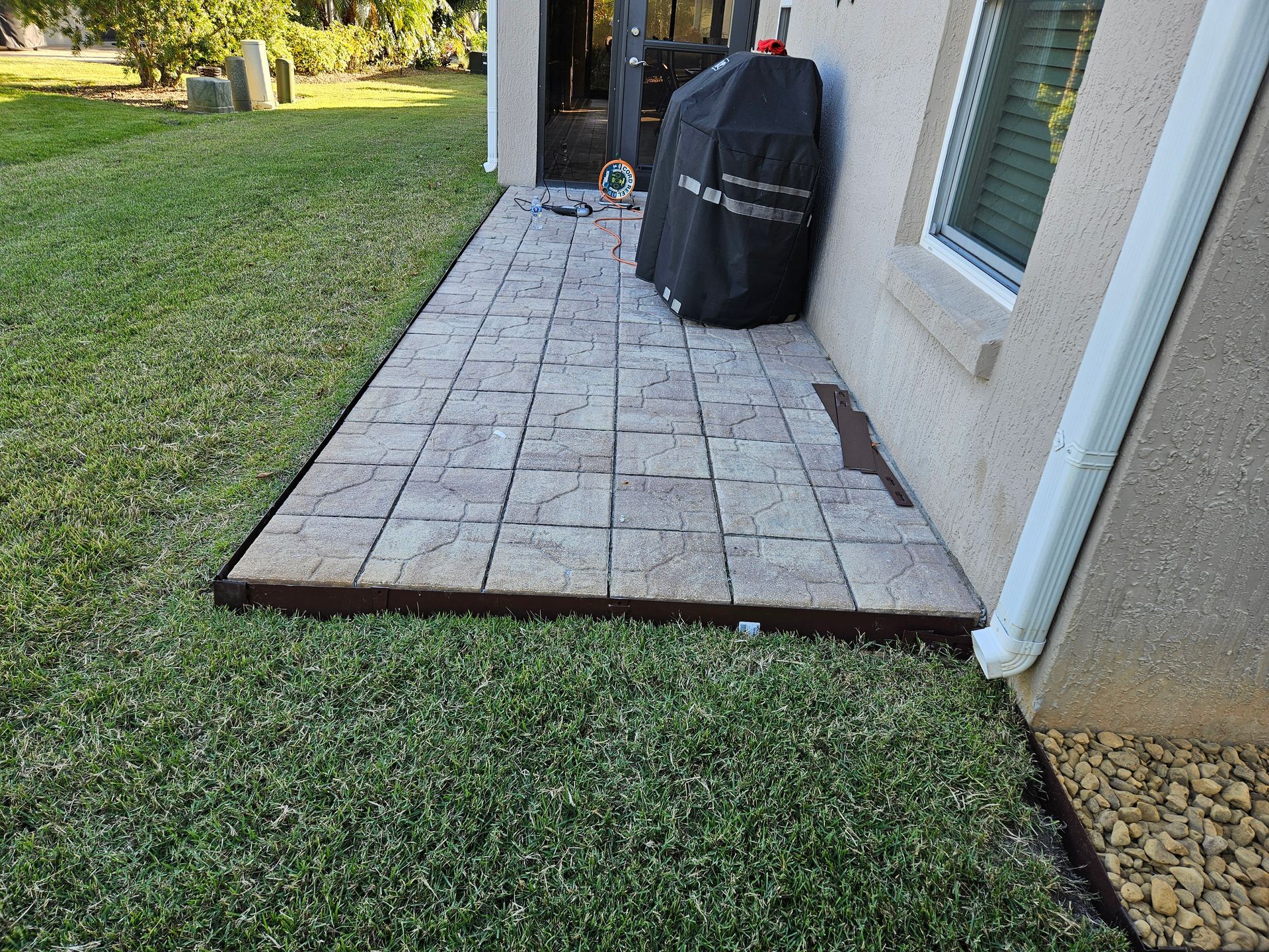 Patio with grill next to a house, bordered by lawn.