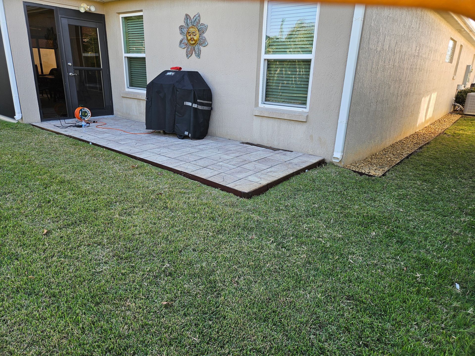 Backyard patio with a grill, bordered by grass and a decorative edge.