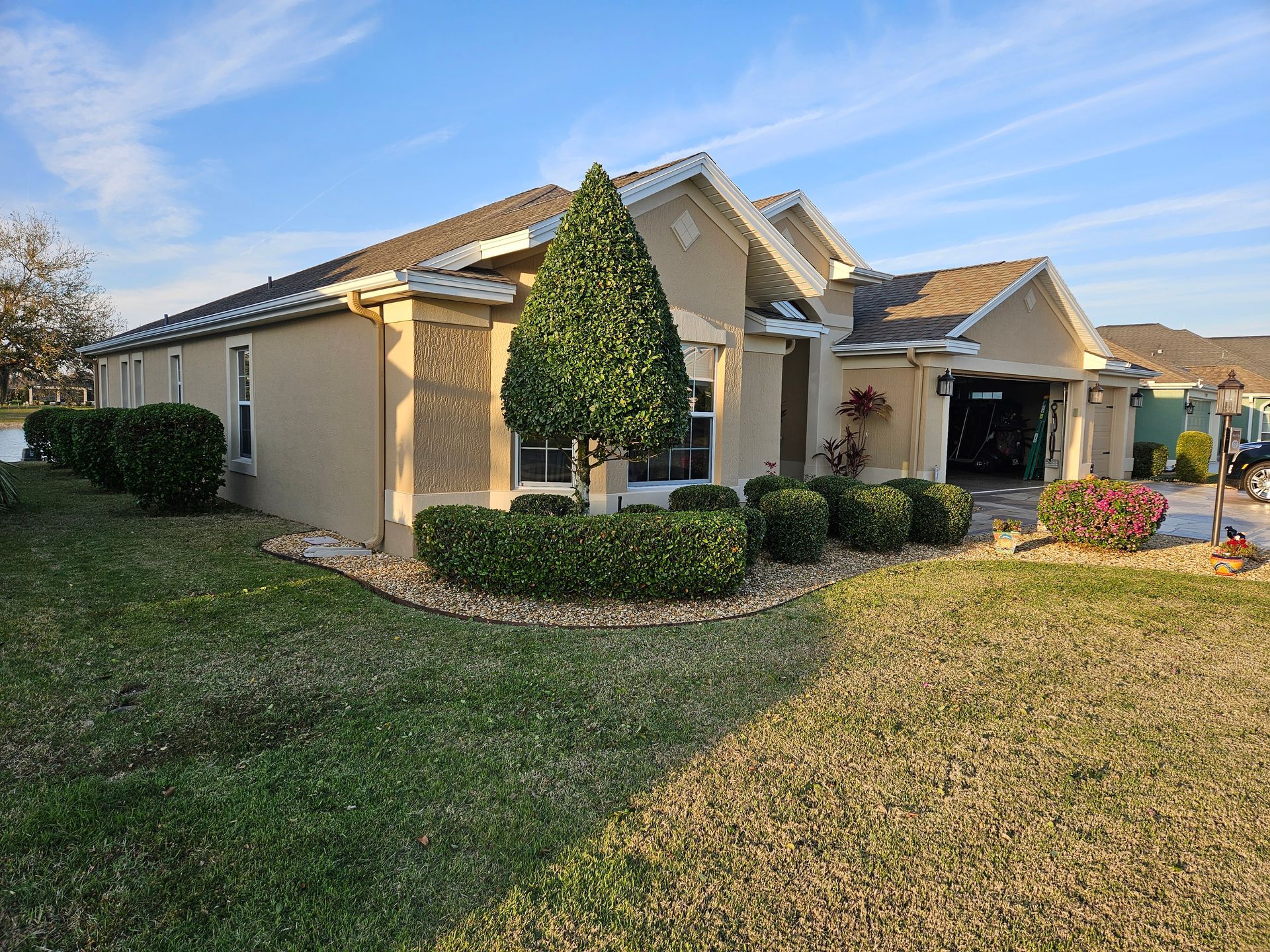 Beige house with well-manicured bushes, a small tree, and a green lawn under a blue sky.