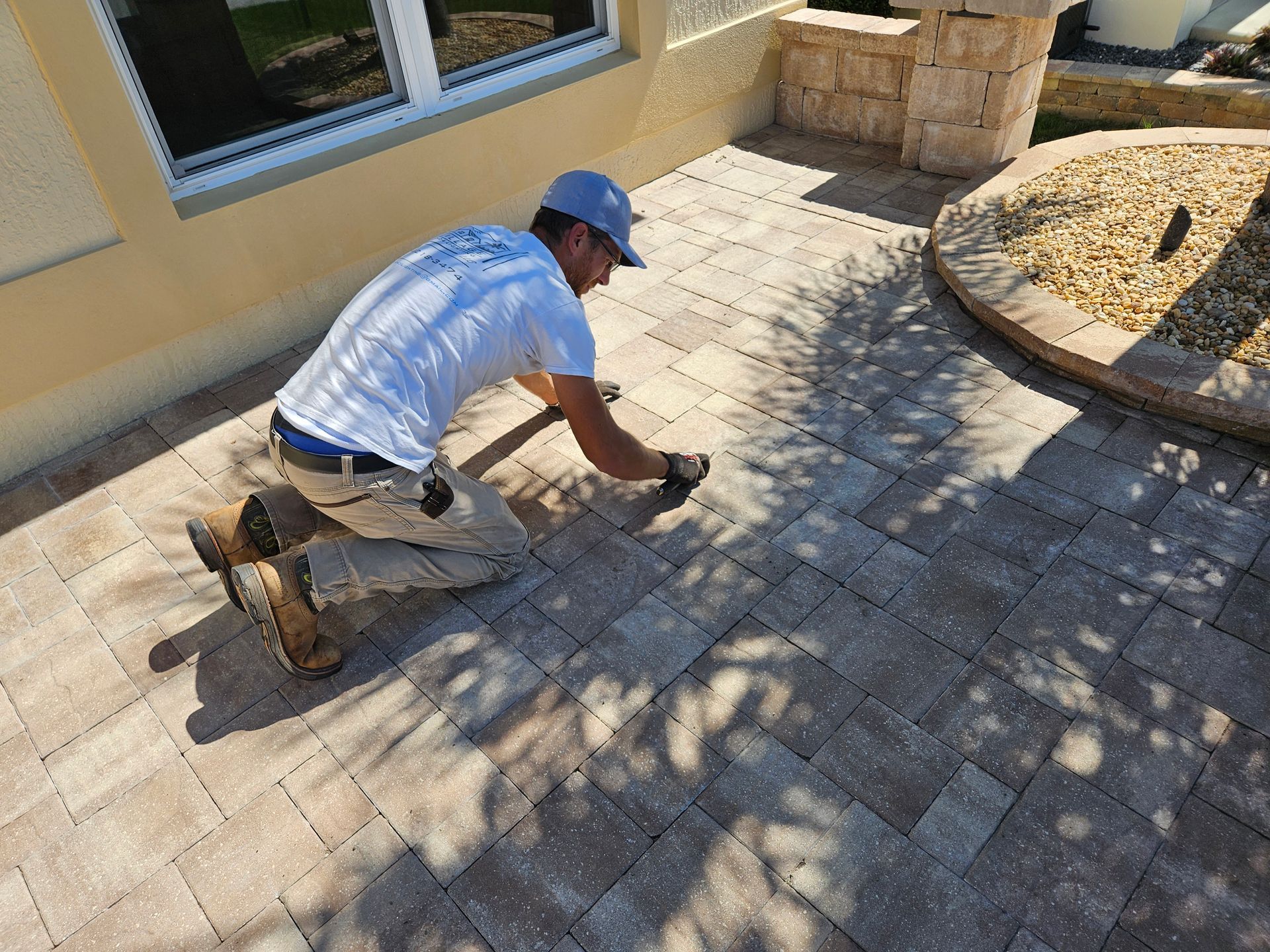 Man kneeling, laying pavers on patio. Tan paving stones, beige stucco wall, blue sky.