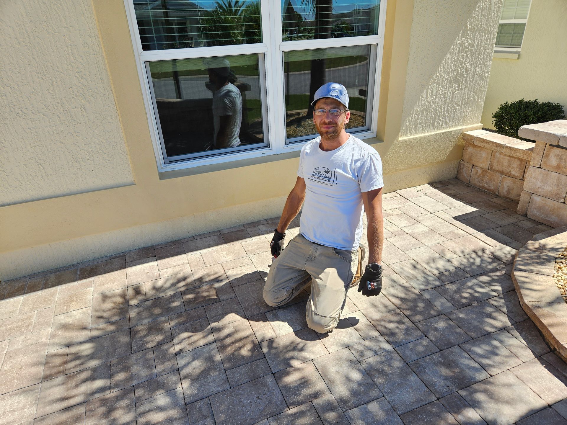 Man kneeling on a brick patio, wearing work clothes and a hat, near a house with a window.