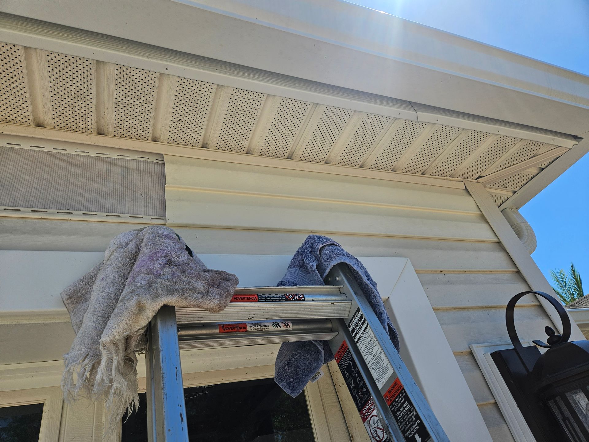 Ladder against a white house, with rags draped over it. Sunlight shines on the siding and soffit.