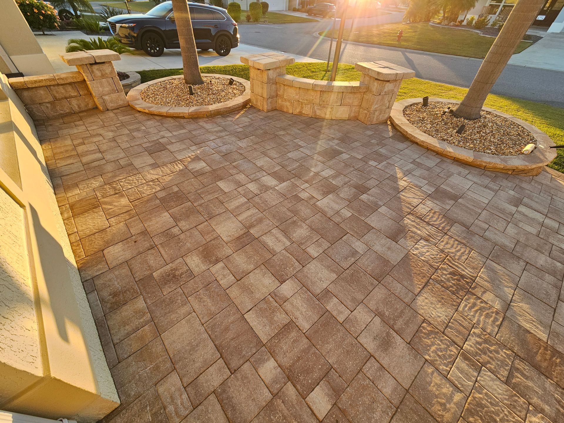 Brick patio with tan pavers, low stone wall, and trees. Sunlight.
