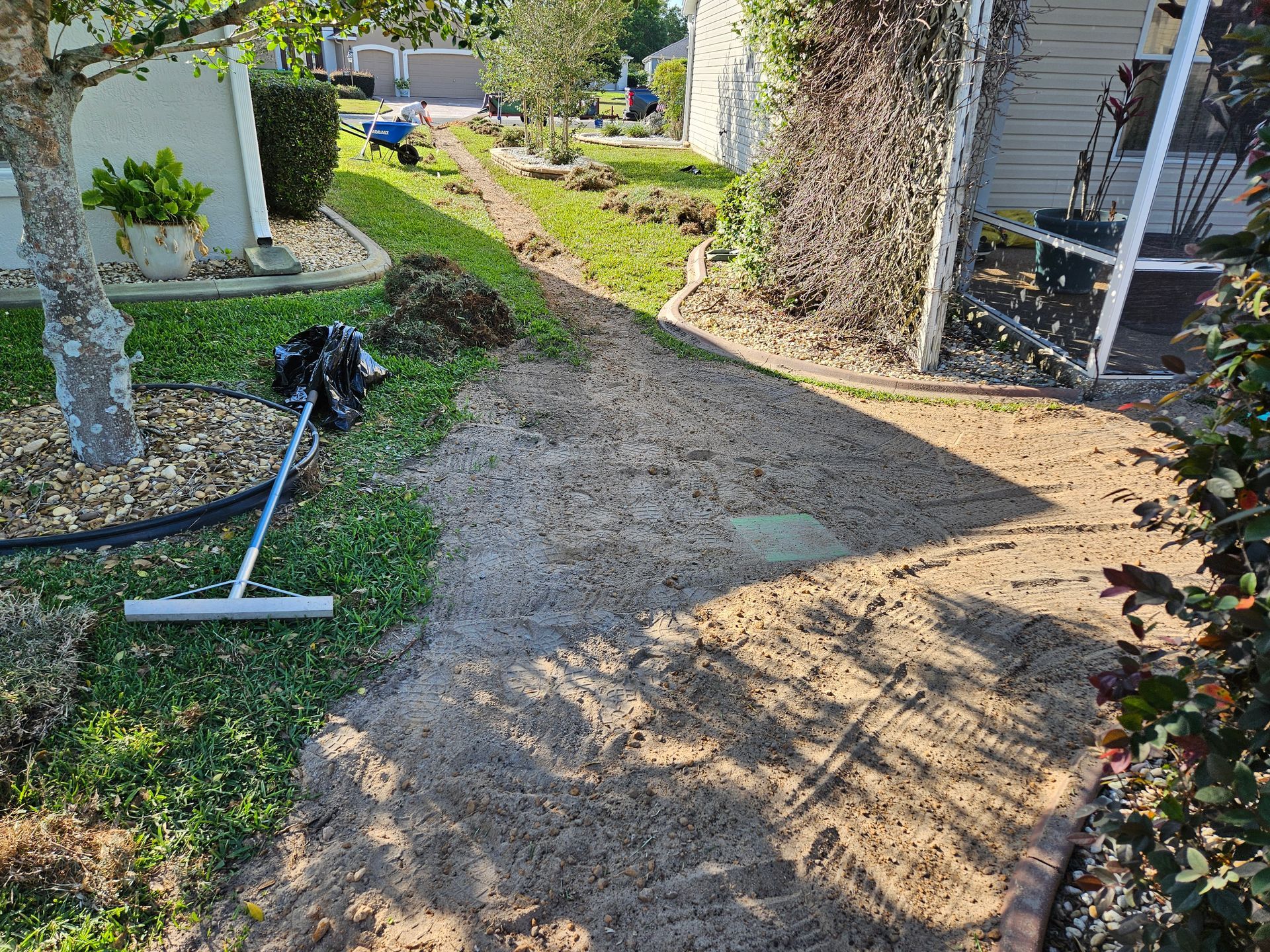 A yard with a trench dug, a rake, and dirt. A home is on the right with trees and a yard.