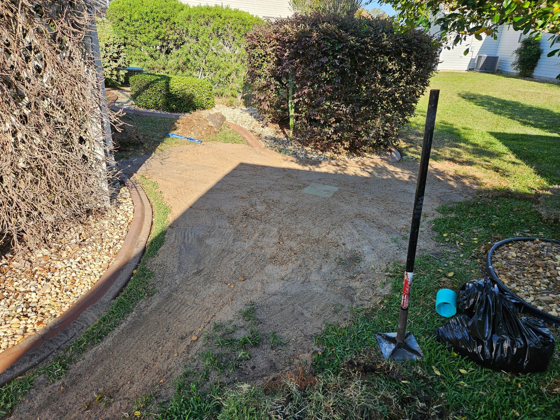 A yard with recently tilled soil, a shovel, bushes, and a black bag on grass.
