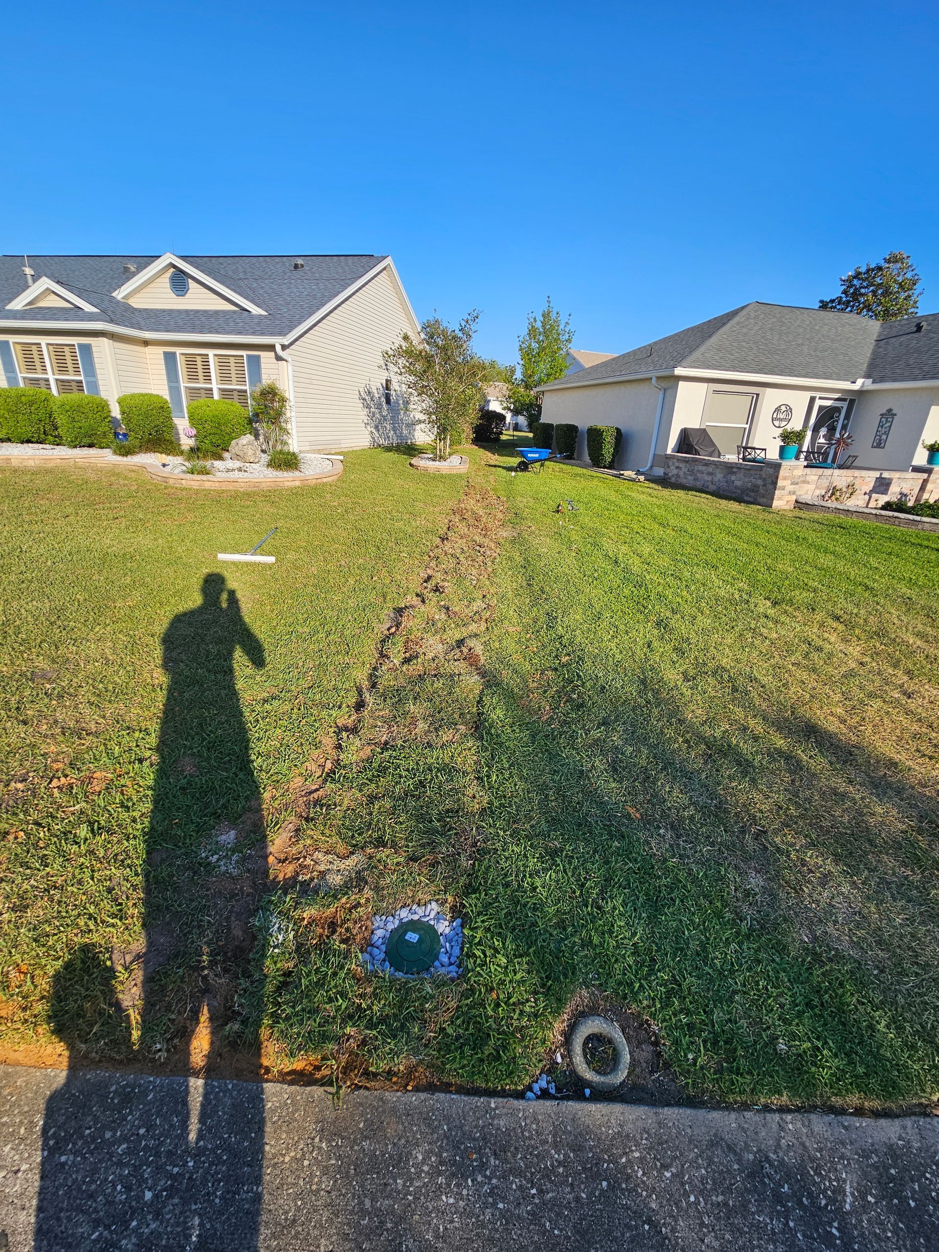 Lawn with patchy grass, houses in the background, person's shadow on the lawn. Sunny day.
