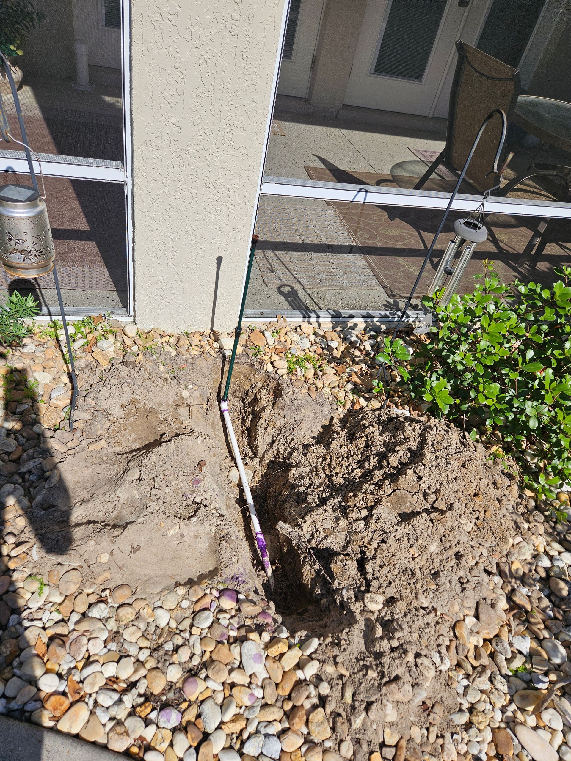 Hole dug in the dirt next to a wall, revealing white and purple pipe. Landscaping stones surround the hole.