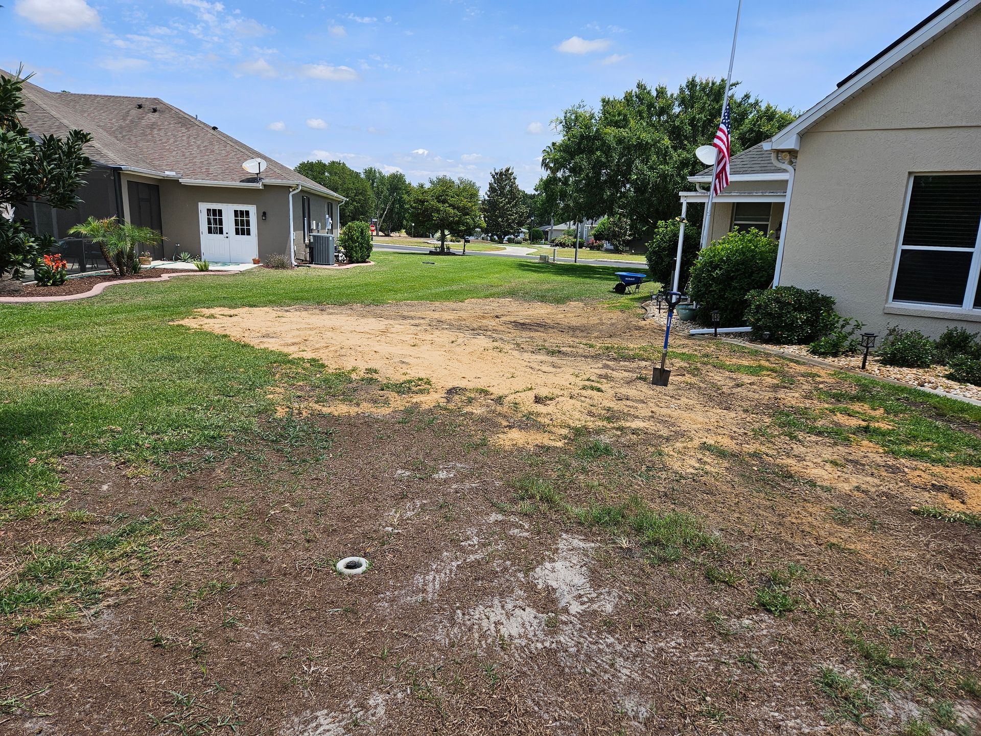 Grassy backyard with patchy brown grass and a bare dirt area between two houses on a sunny day.