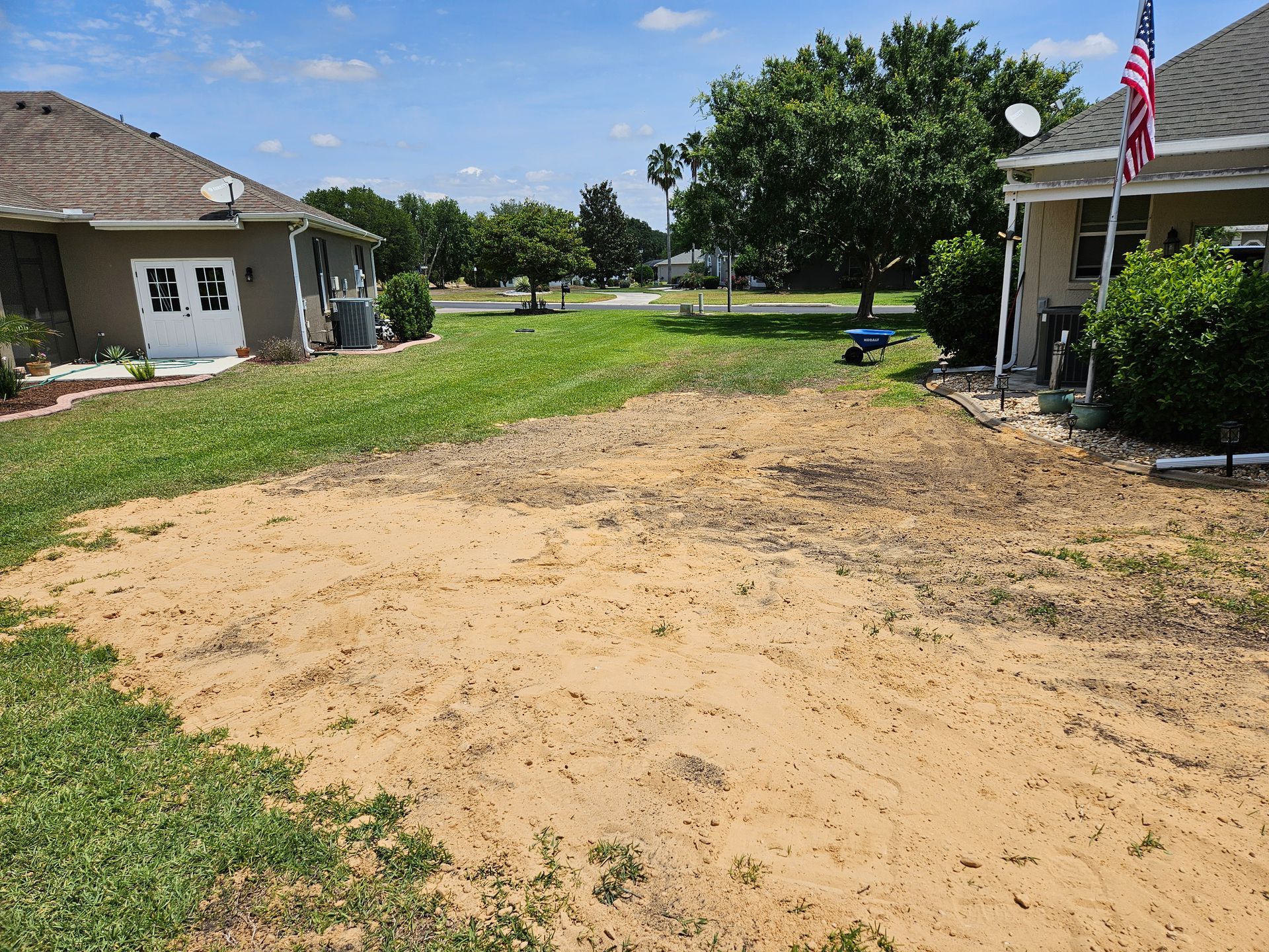 Sandy patch in residential yard between two houses, grassy areas, blue sky.