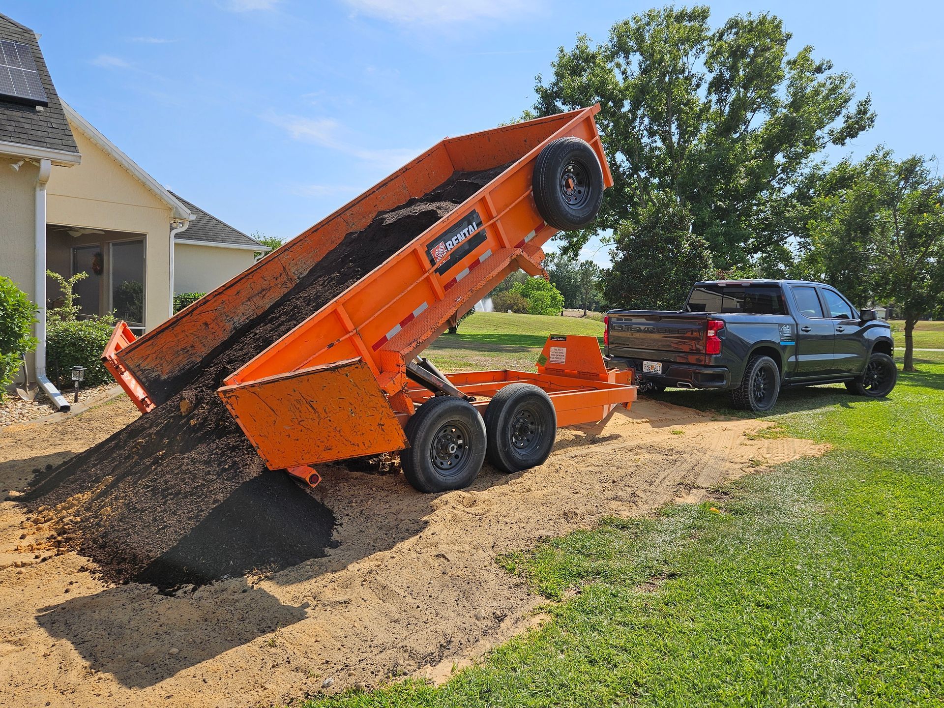 Orange dump trailer unloading asphalt onto a gravel area next to a house, pulled by a black pickup truck.