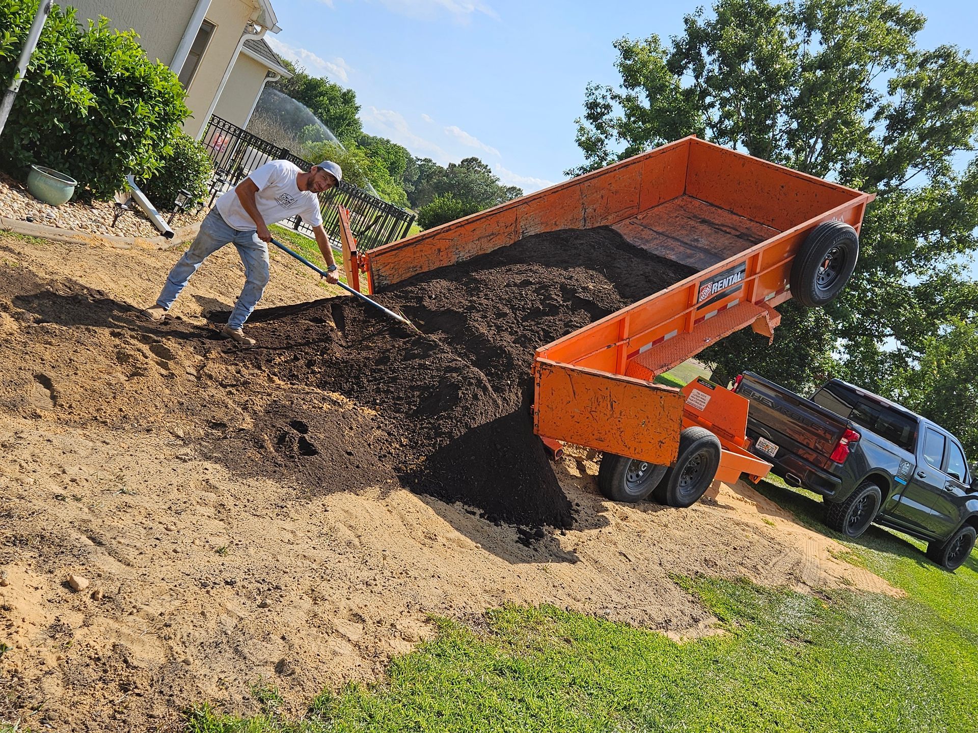 Man shoveling dark mulch from orange trailer onto a sloped yard, parked next to a gray truck.
