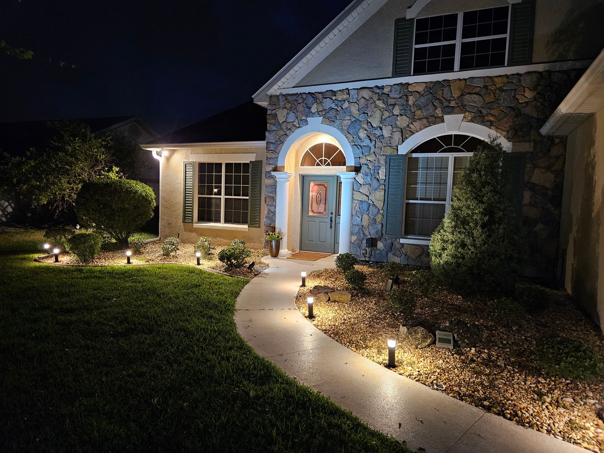 House at night with illuminated walkway, stone facade, and green lawn.