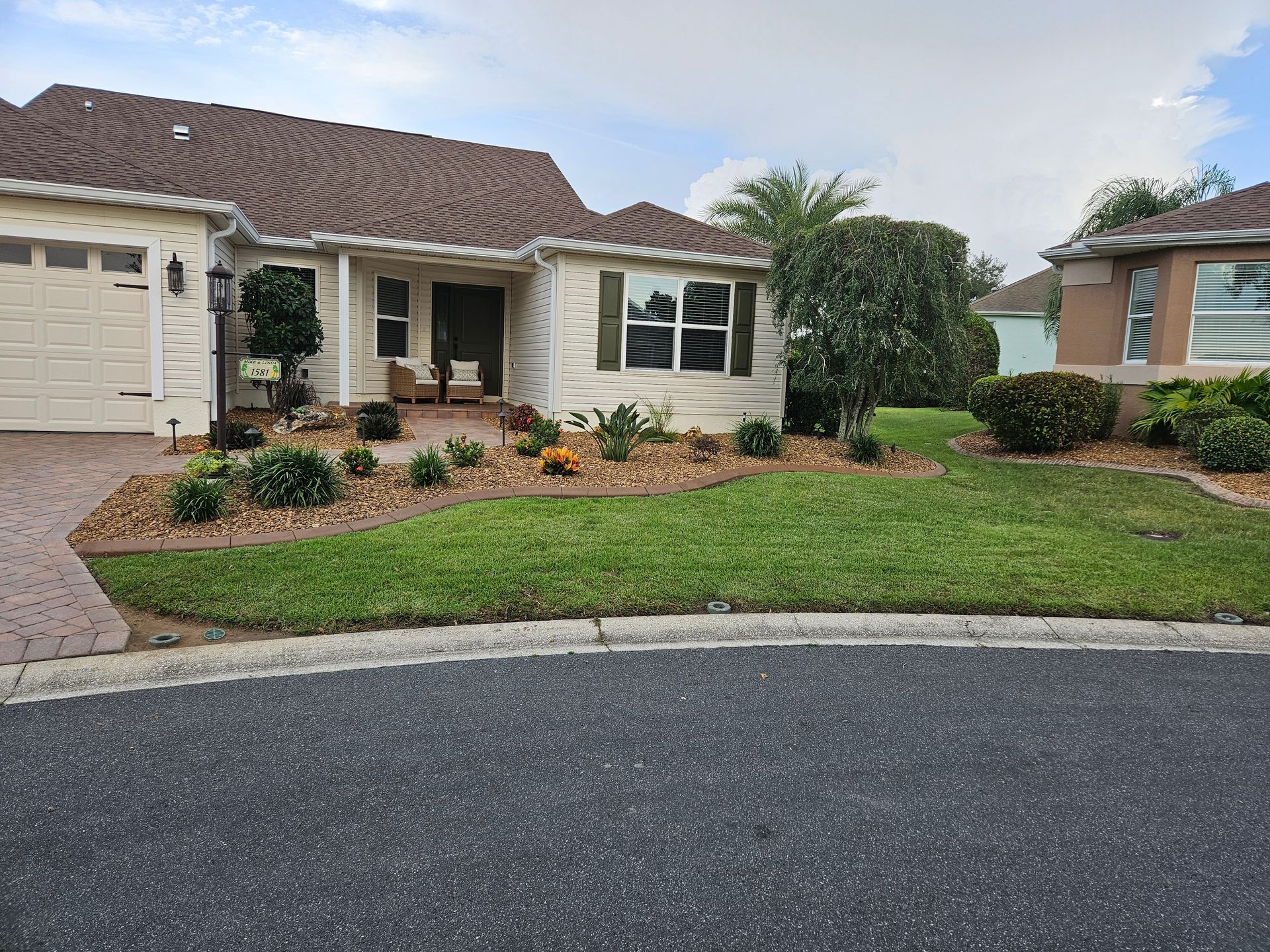 House with tan siding, green lawn, and landscaping. Brown roof and driveway.