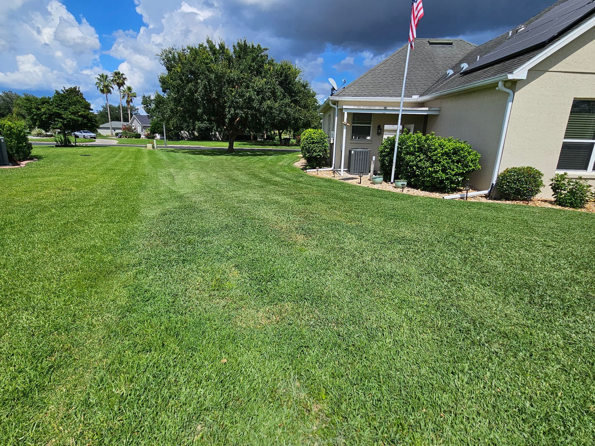 Lush green lawn next to a house with an American flag in the background. Blue sky with clouds.